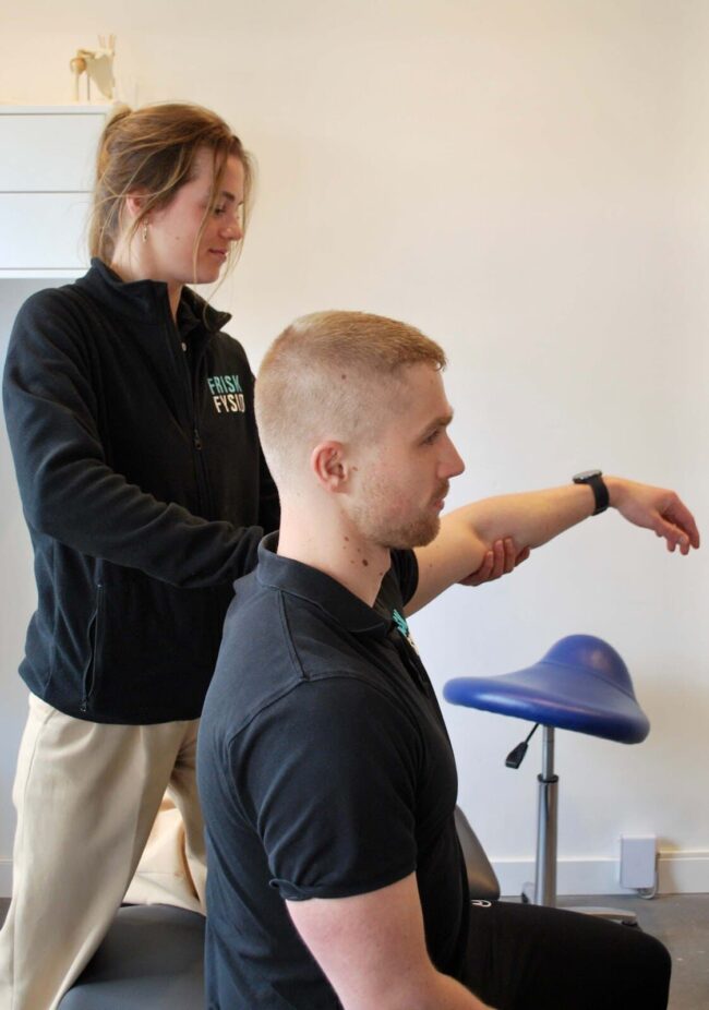 Vrouwelijke fysiotherapeut helpt een man in zwart shirt met oefeningen tegen een peesontsteking in de schouder.