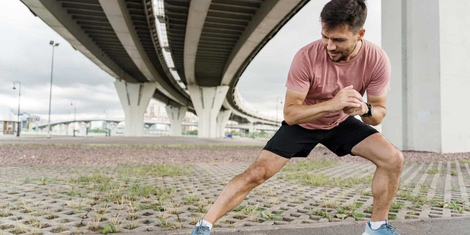 Kramp in de kuiten Man probeert Kramp in de kuiten te voorkomen en doet een oefening buiten onder de viaduct in een roze shirt met sportschoenen