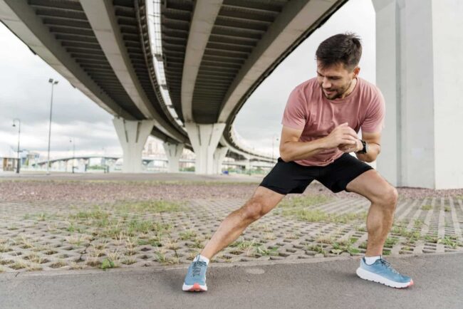 Man probeert Kramp in de kuiten te voorkomen en doet een oefening buiten onder de viaduct in een roze shirt met sportschoenen