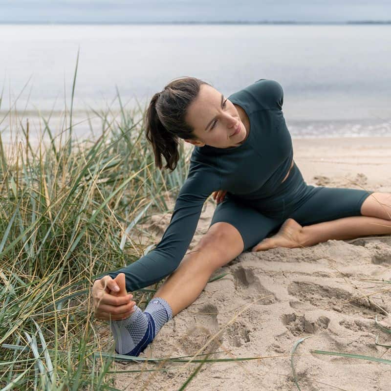 MalleoTrain enkelbrace 10 Vrouw doet op het strand een rek- en stretchesessie bij MalleoTrain enkelbrace, zonovergoten duinen en zee op de achtergrond.