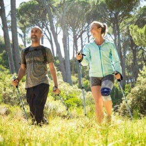 Two hikers wandelen met wandelstokken in een zonovergoten bos, één persoon draagt kniebrace bij knieblessure.