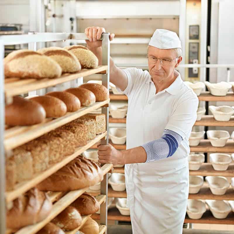 EpiTrain elleboogbrace 6 Franse bakker controleert warme broden uit een rij-ovenrek bij een bakkerij, comfortabel gekleed in wit uniform en hoed.