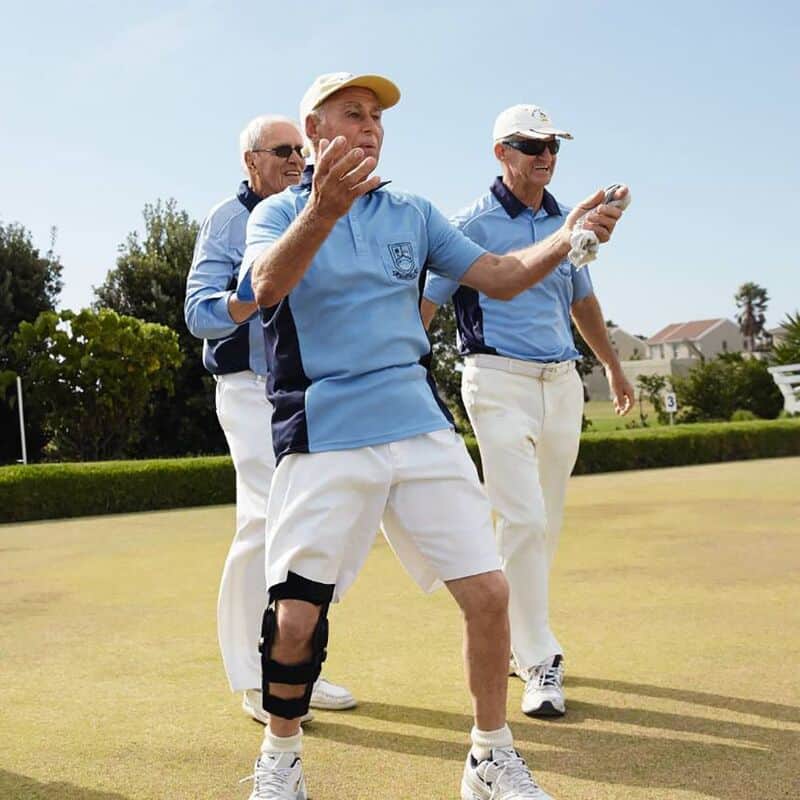 SecuTec® OA 2 Drie oudere mannen in lichtblauwe polo's en witte broeken wandelen samen op een zonnige golfersbaan; één man met knie brace maakt een gebaar terwijl de anderen lachen en plezier hebben.