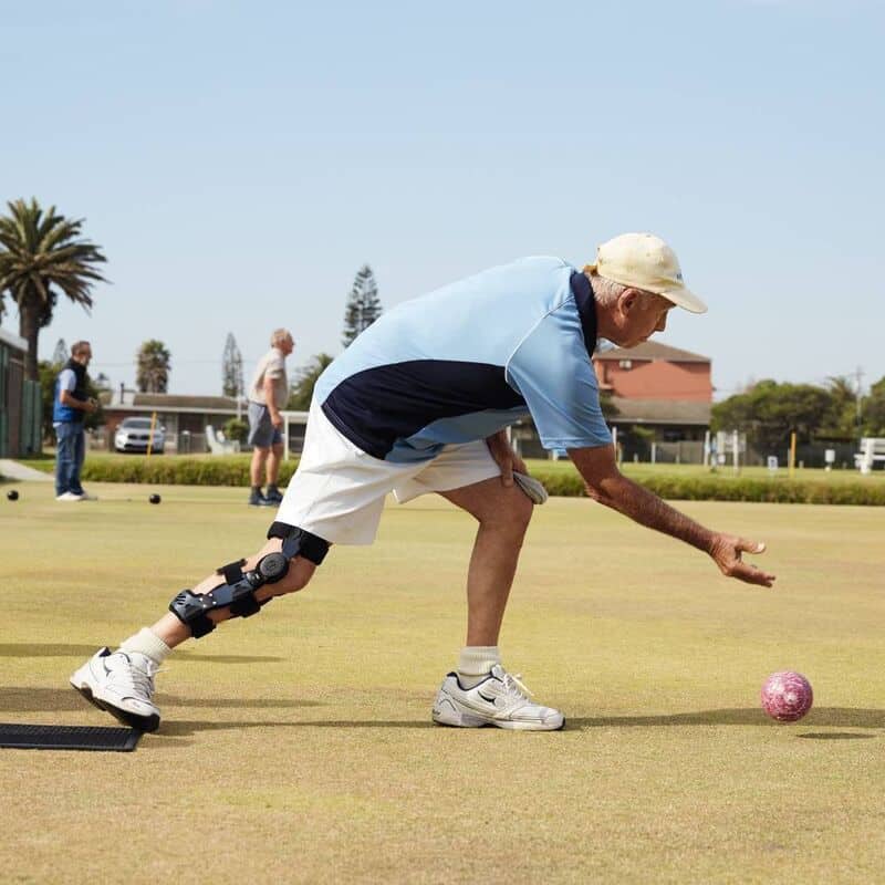 SecuTec® OA 6 Oudere man met knie- en enkelbrace gooit een kogel op een sportveld tijdens een bolavourtocht bij FRISKFYSIO.