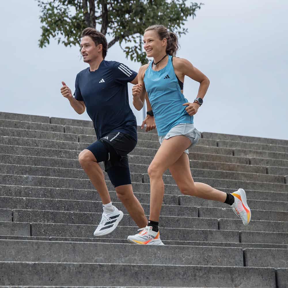 Run Performance Low-Cut Socks 5 Two athletes are running up the stairs together during an outdoor training session: a man in a dark blue shirt and a woman in a light blue top and white shorts; both are wearing breathable Adidas sportswear.
