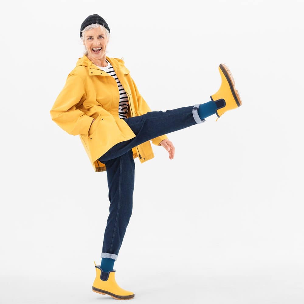 VenoTrain® hero 3 A lively older woman in a yellow coat and wellies performs a high kick as a playful model for FRISKFYSIO, all in a bright studio.