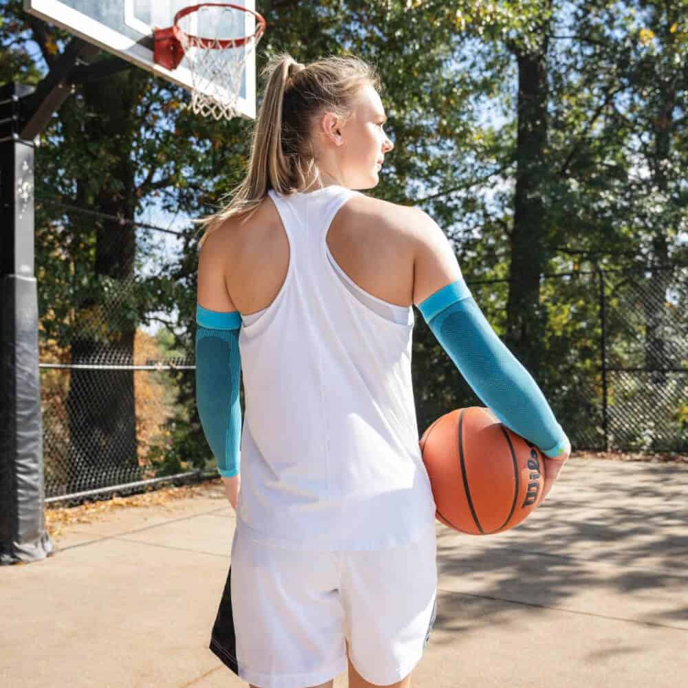 Sports Compression Sleeves Arm 3 Sportende persoon vasthoudt een basketball bij basketbalveld met lange mouwen armbanden en witte sportkleding in zonnige buitenomgeving.