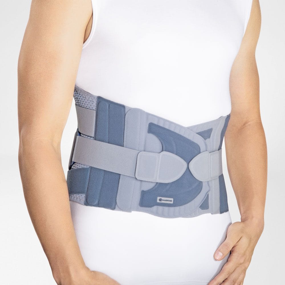 Spinova® Stabi Classic 2 A man is wearing a back brace in shades of blue-grey whilst standing upright, his waist turned slightly to one side to show the back support.
