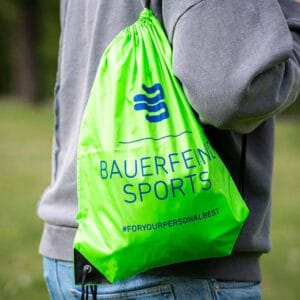 A person carrying a green sports bag bearing the words ‘Bauerenfinl Sports’ and the hashtag ‘#ForYourPersonalBest’ in a park-like setting.