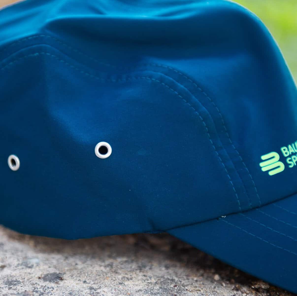 Sports Cap 5 Blauwe sportpet met witte oogringen en lichte stiksels; close-up van een pet met branding op de rand.