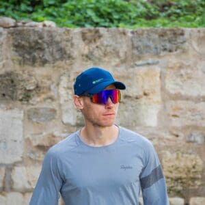An athlete in a light blue sports T-shirt and a blue cap, wearing sunglasses, during an outdoor training session next to a stone wall.