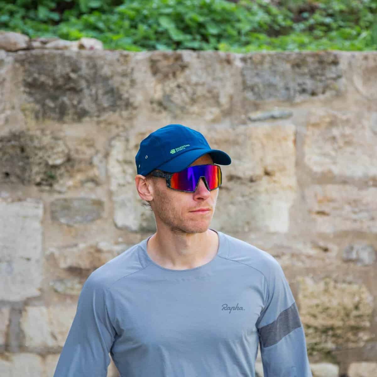 Sports Cap 2 Atleet in lichtblauwe sportt-shirt en blauwe cap met zonnebril tijdens buitentraining naast stenen muur.