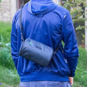A person wearing a blue jacket with a small rucksack-style bag slung over their shoulder, walking outdoors amongst greenery.