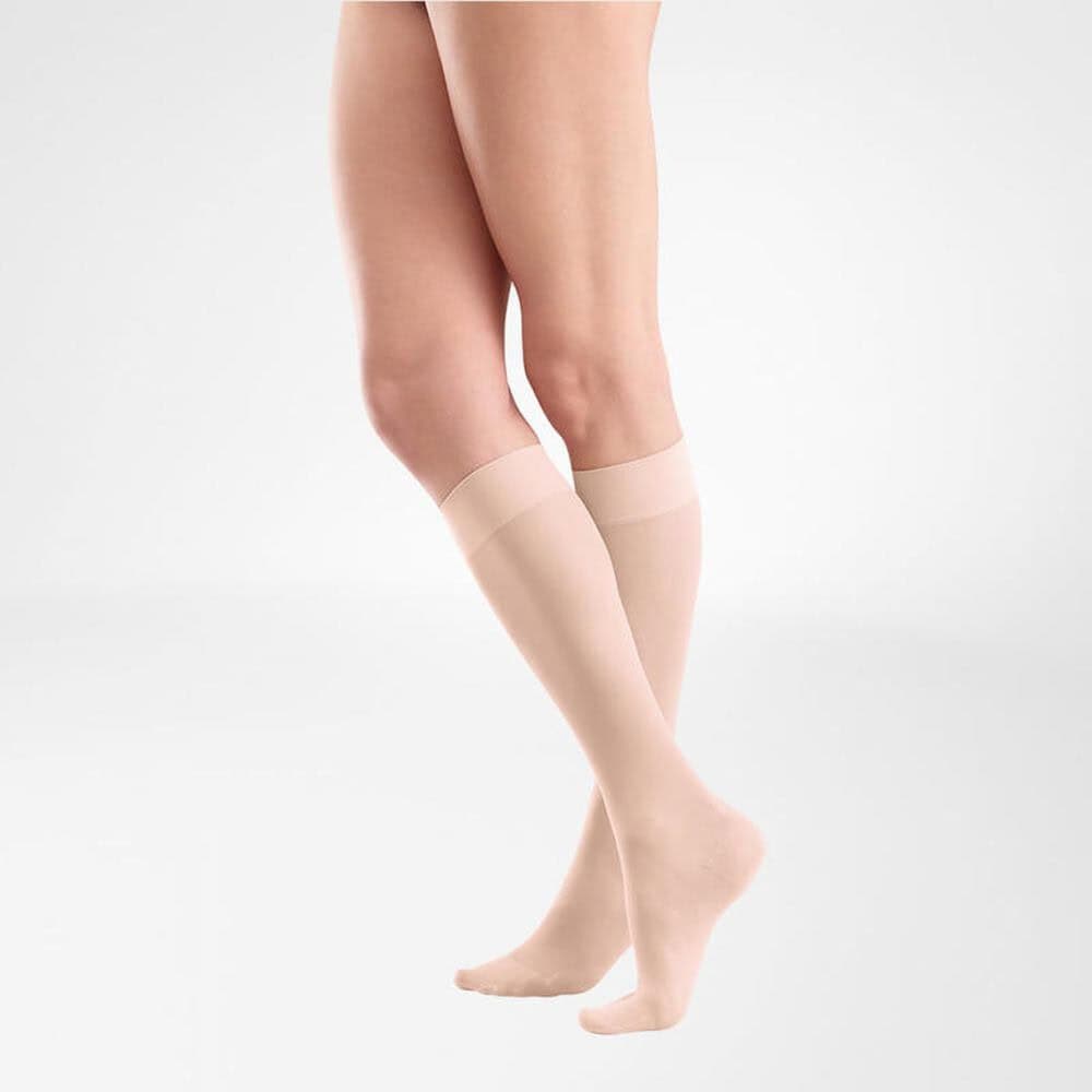 VenoTrain® act SHEER ELEGANCE AD 7 Vrouwelijke benen met naadloze kuit- en enkelkousen in huidkleur, staand tegen een lichte achtergrond.