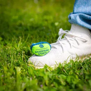 A foot wearing white trainers on a grassy patch, with a striking green and yellow ball from Bauerfeind Sports next to the trainer.