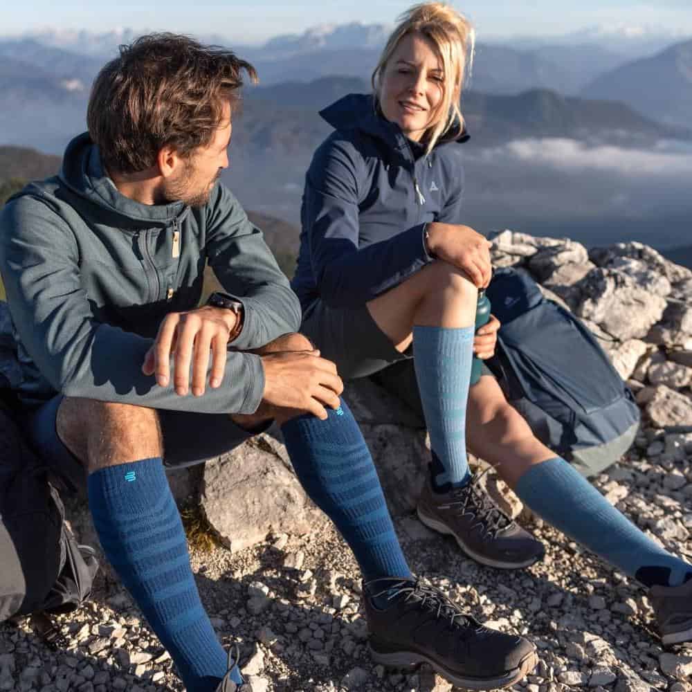Outdoor Merino Compression Socks 6 Two athletes take a break during a mountain walk on a rocky peak with views of the mountains and a clear sky.
