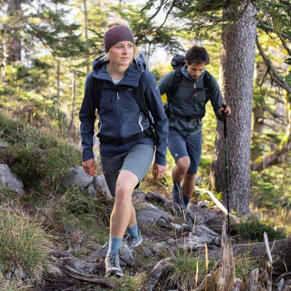 Outdoor Merino Mid Cut Socks 5 Hardlopers in een bosrijke omgeving tijdens een hike, vrouw voorop met hoofddeksel en rugzak, wandelstokken en stevige wandelschoenen.