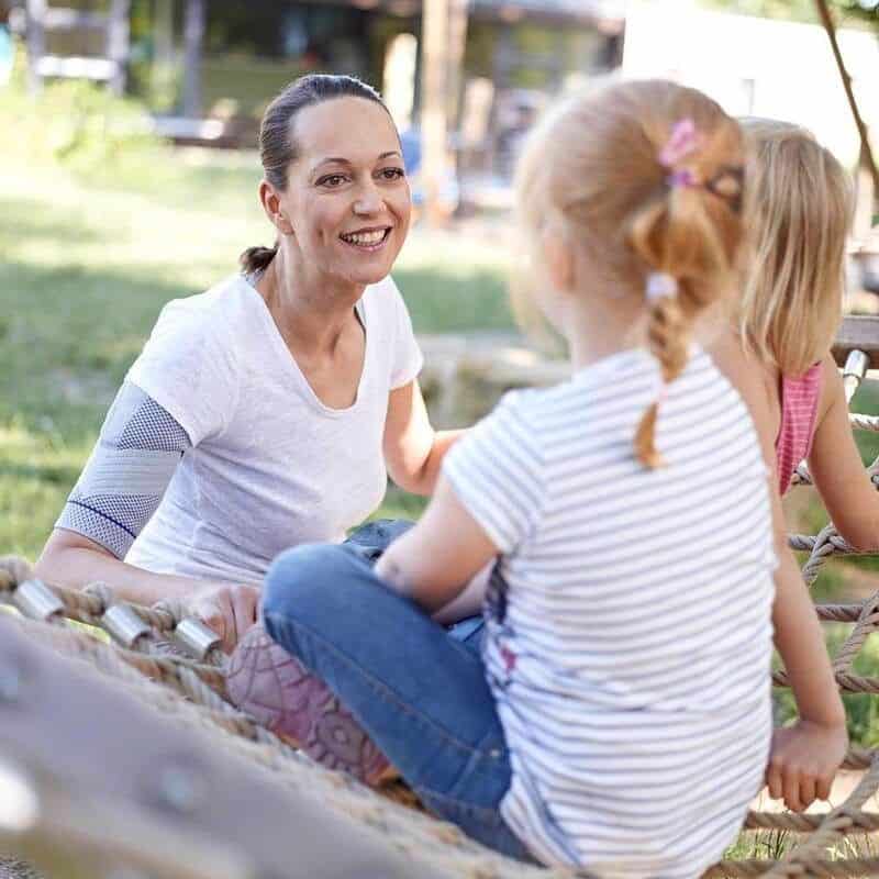 OmoTrain schouderbrace 6 Vrouw met een schouderbrace op lamping outdoor training, glimlachend terwijl ze met kinderen praat.