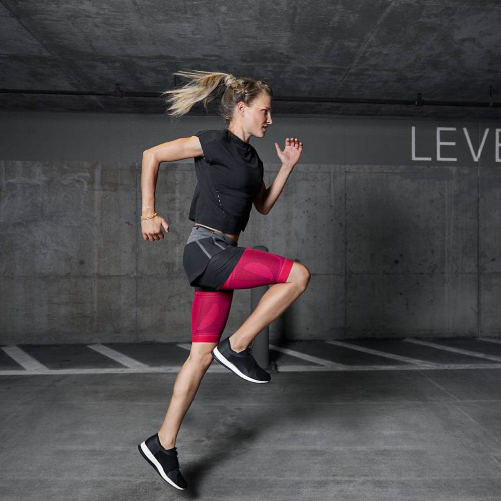 Sports Compression Sleeves Upper Leg 3 Actieve sportvrouw rent snel vooruit in een industriële trainingsruimte met halogeenverlichting en betonmuur, gericht op krachttraining en conditie.