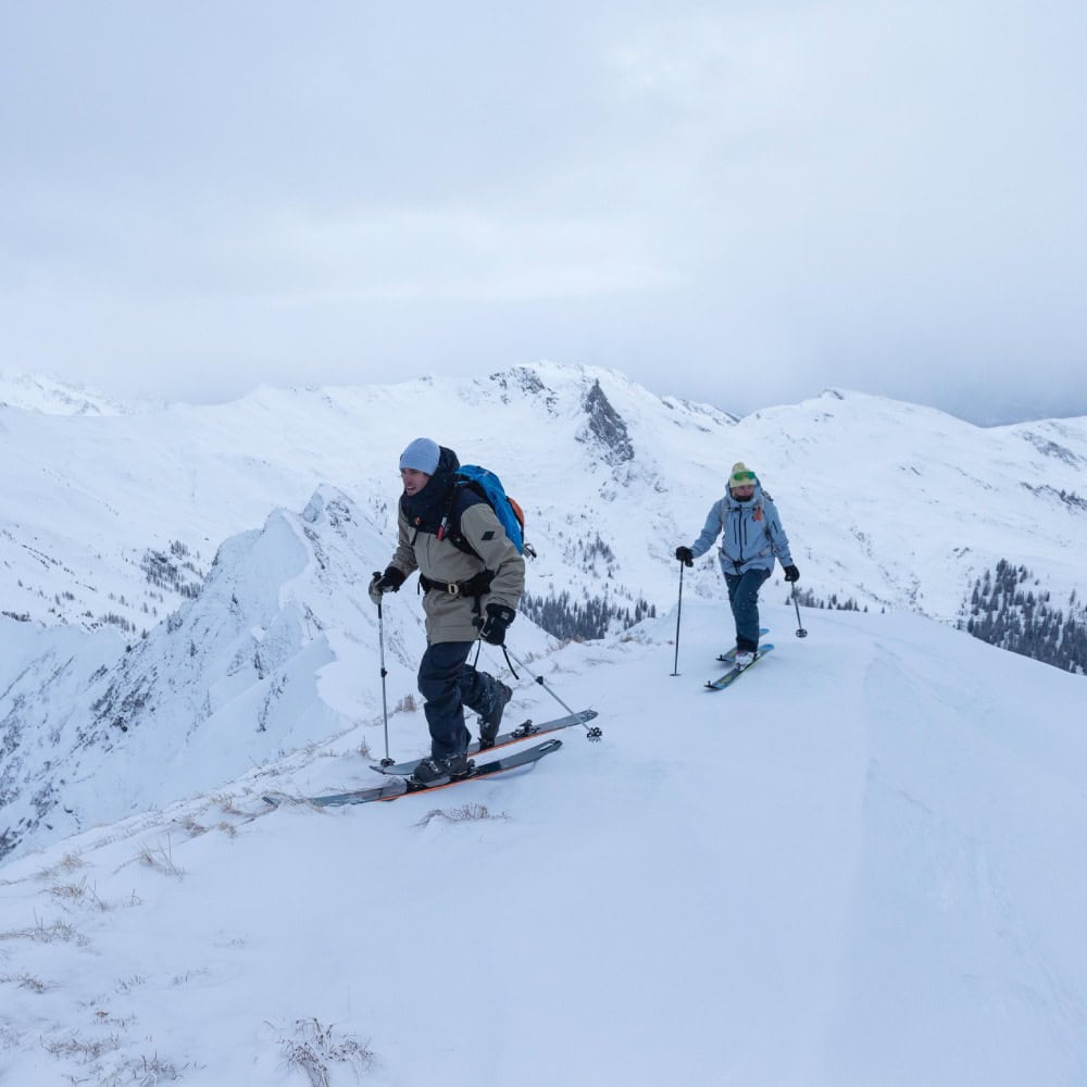 Ski Touring Compression Socks 6 Two skiers are climbing a snow-covered ridge under a cloudy sky in a winter landscape, carrying walking sticks and rucksacks.