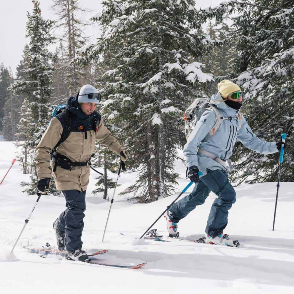 Ski Touring Compression Socks 5 Two skiers skiing uphill through a snow-covered pine forest, carrying their equipment and ski poles.