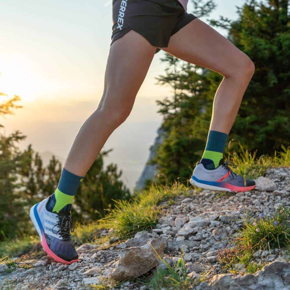 Trail Run Mid-Cut Socks 5 A runner on a mountain path wearing trail running shoes, against a sunny backdrop and a green landscape.