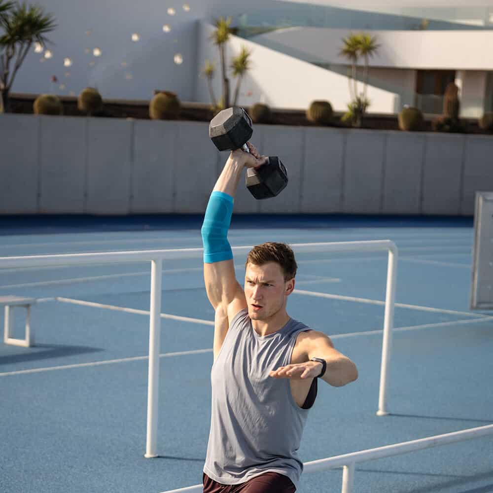 Sports Compression Elbow Support 5 Man doet een eenarmige zwaardere dumbbell jerk boven een atletiekbaan, gericht op krachttraining en fysiotherapie bij FRISKFYSIO NL.