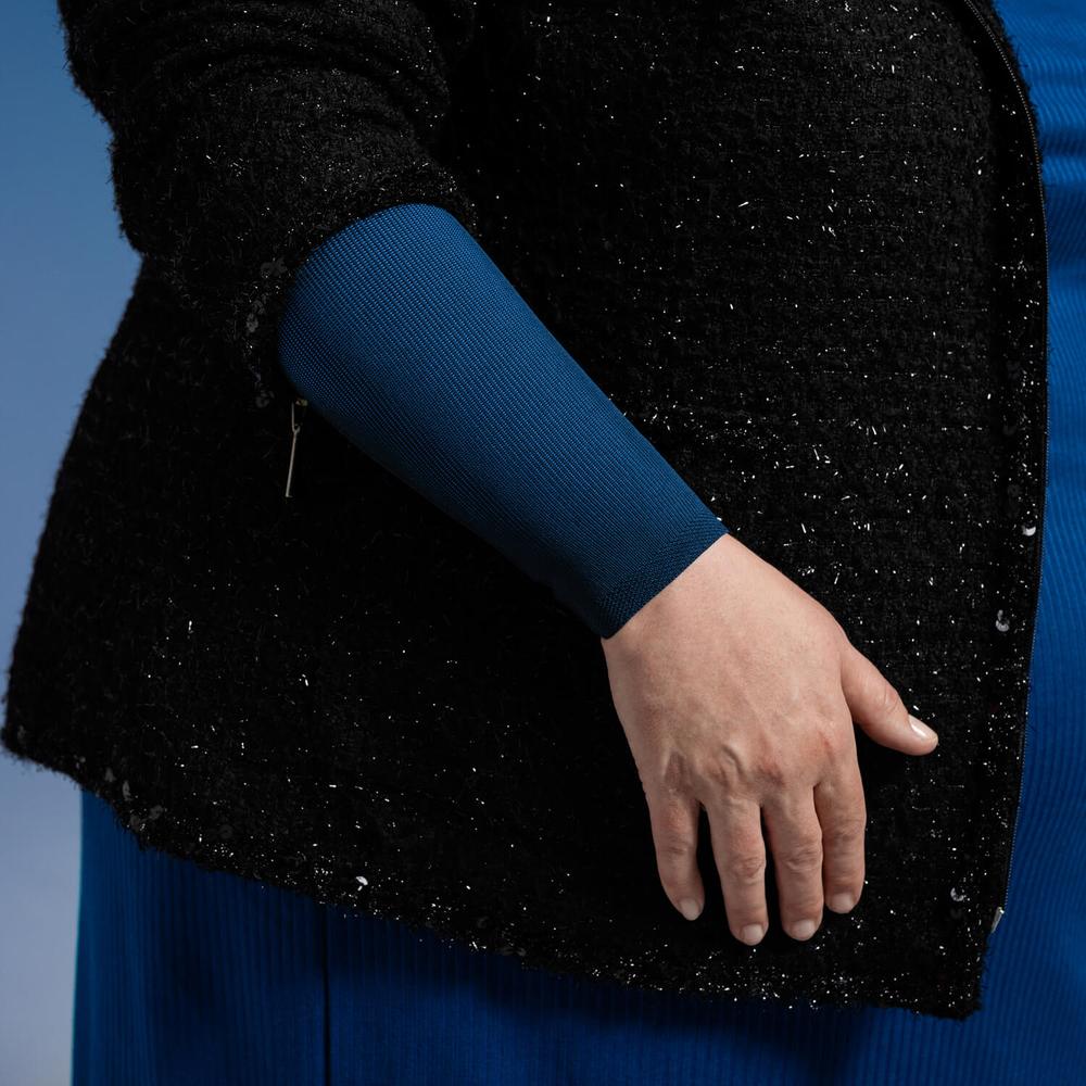 VenoTrain® curaflow armkousen 12 Man vrouw draag een donker geblokte jas met een ultramarijnblauwe mouw zichtbaar, vastgelegd tegen een heldere blauwe achtergrond.
