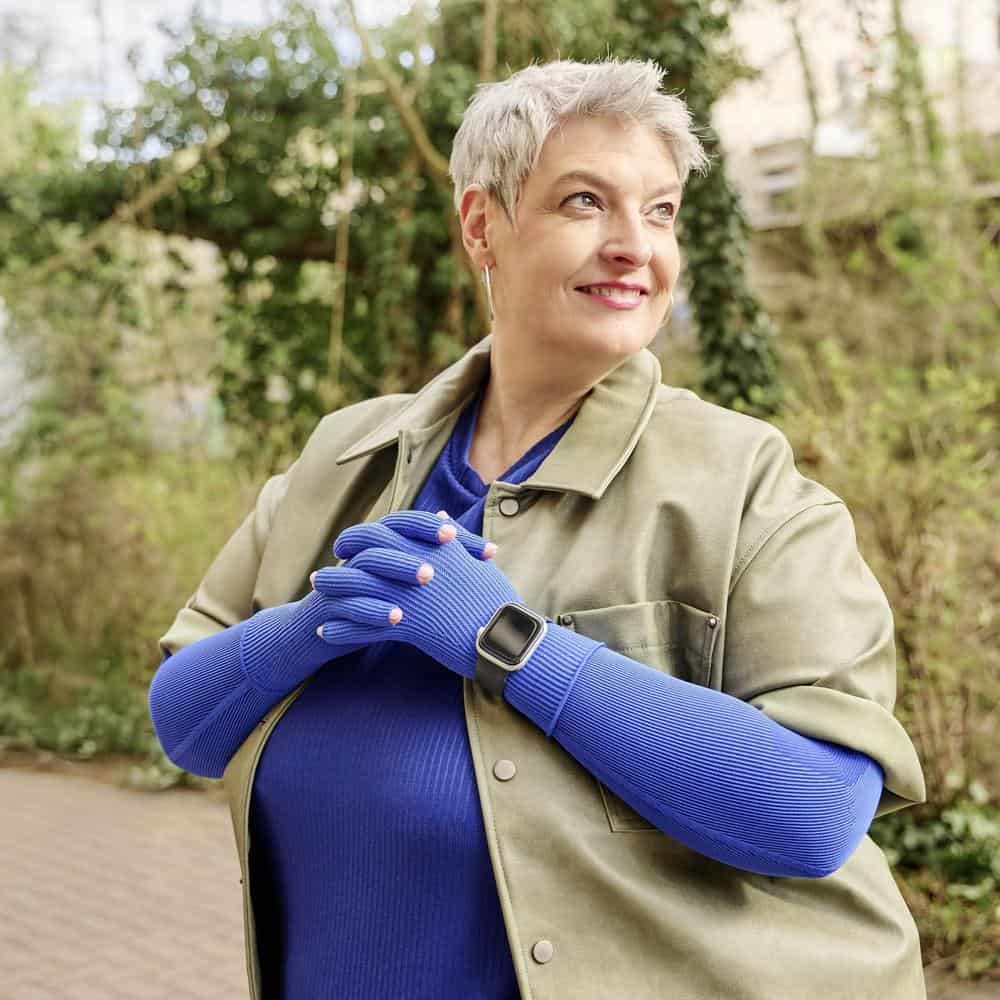VenoTrain® curaflow armkousen 13 Frisse oudere vrouw met korte grijze haren draagt een groene jas en blauwe handschoenen, staand in een tuin en glimlachend naar de lucht.